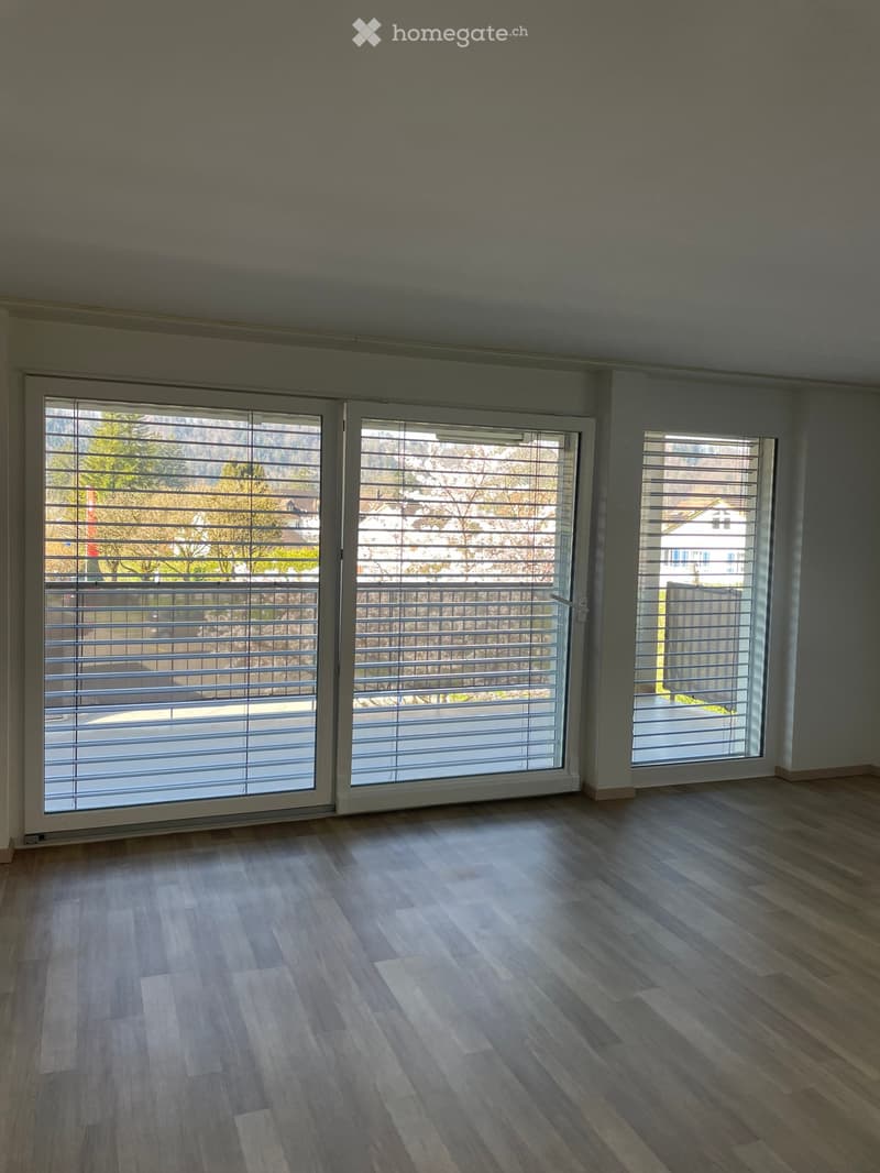 Empty room with wooden flooring and large windows with blinds, offering a view of the outdoors.