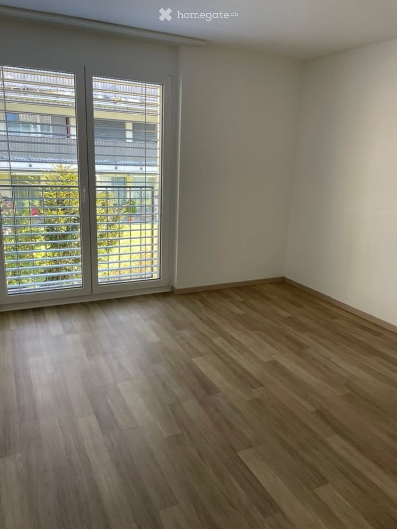 Empty room with wooden flooring and large windows with blinds, leading to a balcony area.