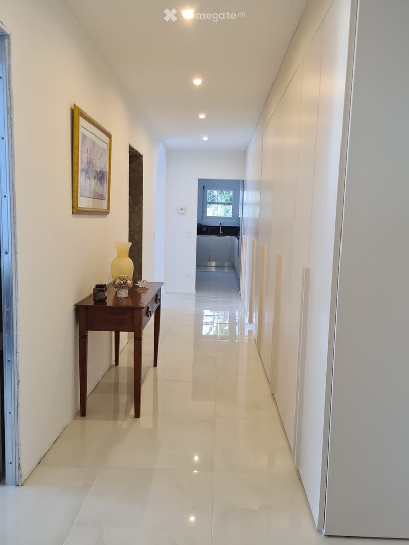 Bright hallway with white walls, polished tile floor, small wooden table with vase and flowers, and a framed picture on the wall. The hallway leads to a kitchen area visible at the end.
