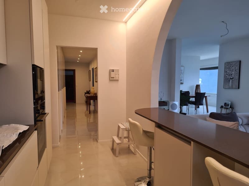 Modern hallway with white walls, recessed lighting, and a polished tile floor. Features a built-in oven, a small bar area with stools, and a view into a dining area with a table and chairs.