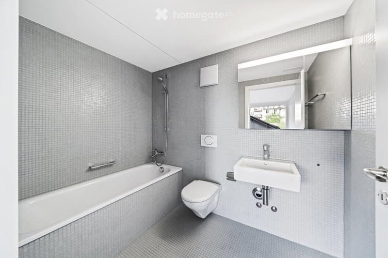 Modern bathroom with grey tiled walls and floor, featuring a bathtub, toilet, sink, and large mirror with built-in storage.