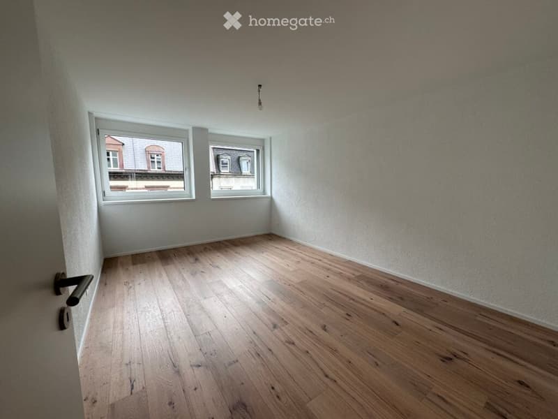 Empty room with wooden flooring, white walls, and two windows offering a view of neighboring buildings.