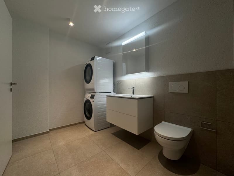 Modern bathroom with a white sink, toilet, mirror, and stacked washing machine and dryer. The walls are tiled in neutral tones, and the floor is tiled with beige tiles.