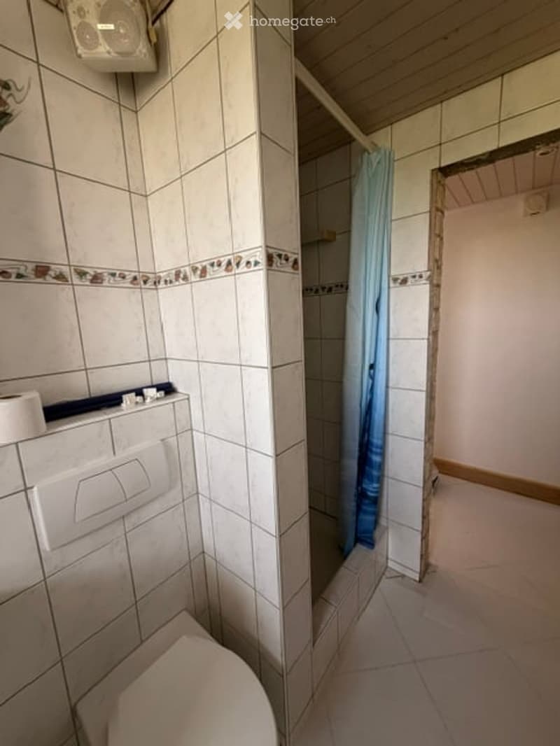 A small bathroom with white tiled walls and floor, a toilet, a shower with a blue curtain, and a toilet paper holder on the wall.