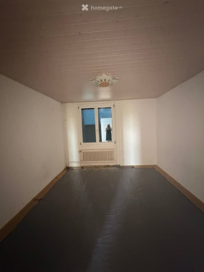 Empty room with white walls, dark flooring, a single window with blinds, a radiator, and a ceiling light fixture.