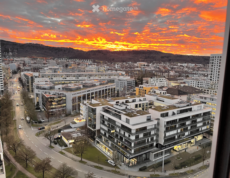 A panoramic view of a cityscape at sunset, featuring modern buildings, roads, and a distant mountain range.