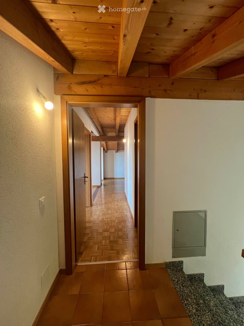 A well-lit hallway with wooden beams on the ceiling, wooden herringbone flooring, and a tiled section at the entrance. The walls are painted in a light cream color, and there is a light fixture on the left wall.