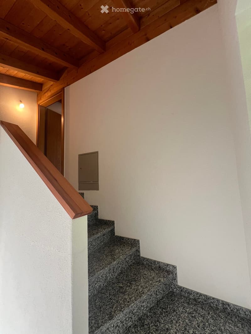 Wooden staircase with a wooden handrail, gray tiled steps, and white walls. A small light fixture is mounted on the wall, and a wooden door is visible at the top of the stairs.