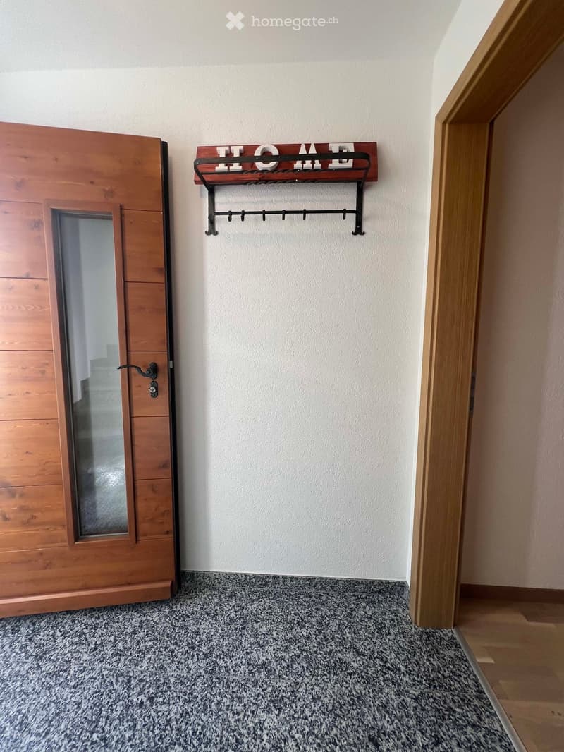 Empty hallway with a wooden door on the left, a coat rack with a "HOME" sign on the wall, and a speckled tile floor.