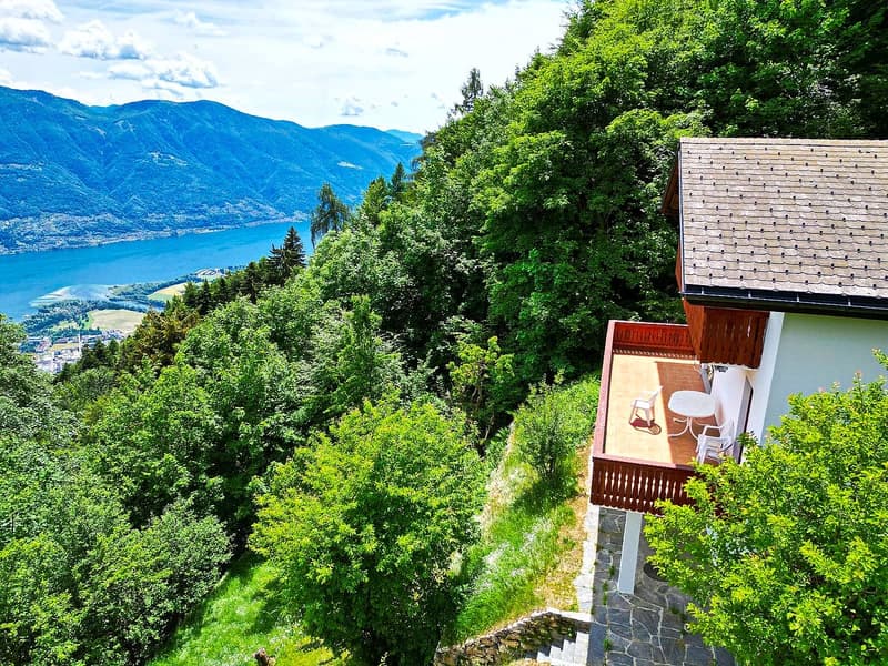 A house with a balcony overlooking a scenic lake and mountain landscape, surrounded by lush greenery.