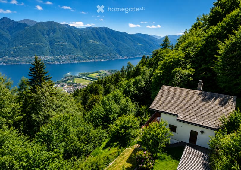 A small white house with a tiled roof, surrounded by lush green trees and shrubs, overlooking a serene lake and distant mountains.