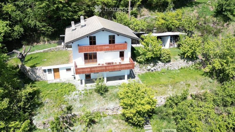 Two-story house with light blue walls, wooden balcony, stone foundation, and surrounding greenery.