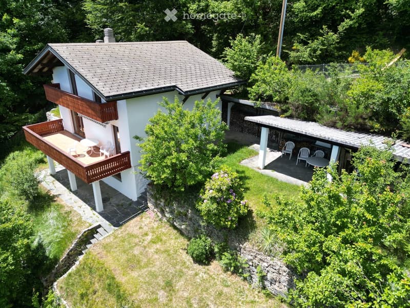 A two-story house with a gray roof and white walls, featuring a wooden balcony and outdoor seating area surrounded by greenery.