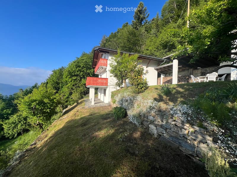 A two-story house with white walls, red balconies, and a stone retaining wall, surrounded by lush greenery and trees.