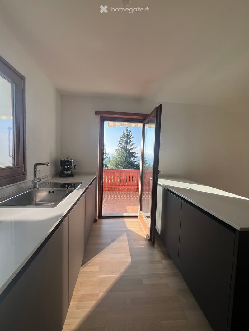 Modern kitchen with white countertops, stainless steel sink, and a coffee maker on the counter. An open sliding door leads to a balcony with a view of trees.