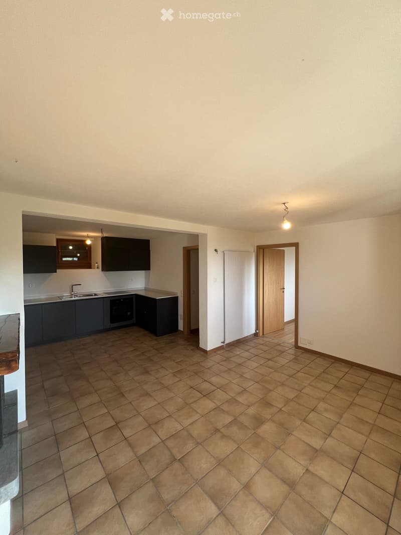 Empty room with tiled floor and beige walls, featuring a doorway leading to a kitchen area with cabinets and appliances.