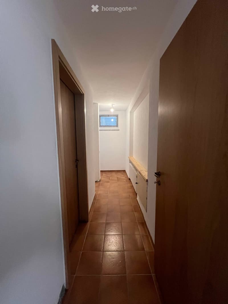 A narrow hallway with tiled flooring, white walls, and a small window at the far end. There are two doors on either side and a small built-in shelf unit on the right wall.