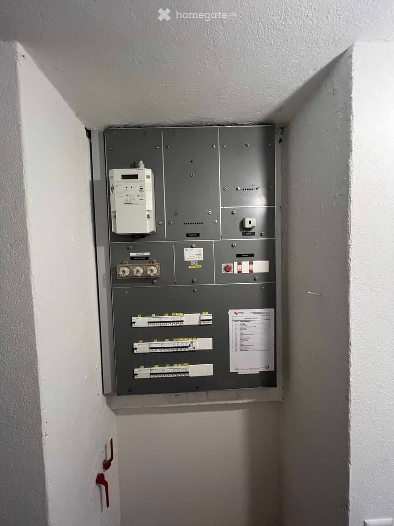 Electrical panel mounted on a white wall in a narrow hallway, featuring circuit breakers, switches, and a digital display unit.