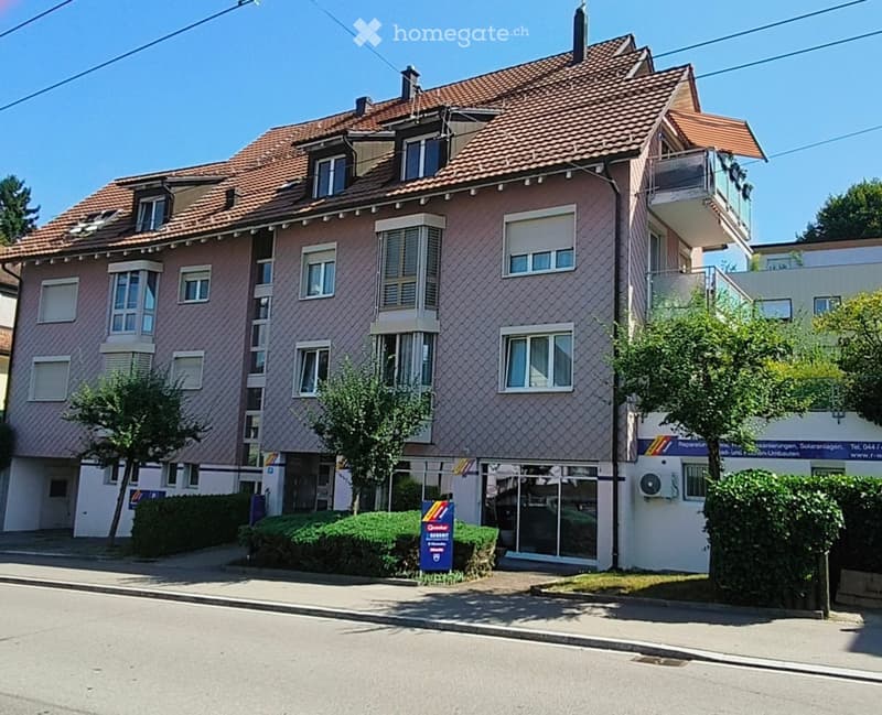 Two-story residential building with pink facade, multiple windows, and a tiled roof. Features a balcony on the upper floor and a small garden area in front.