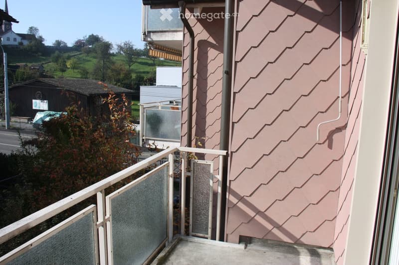 Balcony with metal railing and glass panels, overlooking a residential area with houses, trees, and a clear sky.