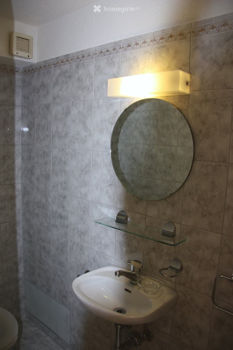 Bathroom with a white sink, chrome faucet, round mirror, glass shelf, and a wall-mounted light fixture. The walls are tiled with a decorative border near the ceiling.