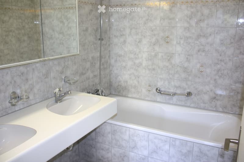 Bathroom with a double sink, bathtub, and a large mirror above the sink. The walls are tiled with a marble-like pattern.