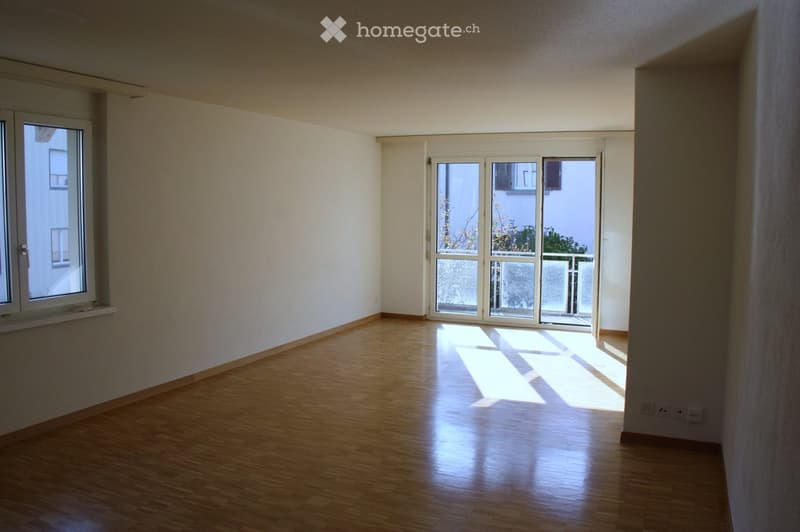 Empty room with wooden flooring and large windows, allowing natural light to fill the space. The room features a sliding glass door leading to a balcony area.