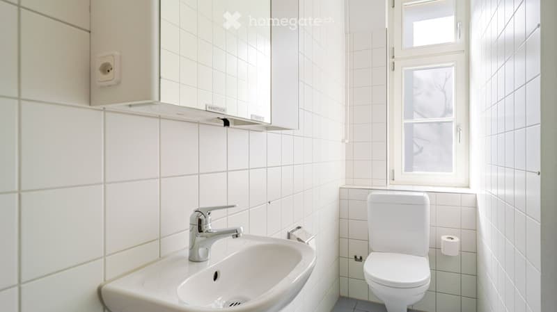 Small bathroom with white tiled walls and floor, featuring a sink with a modern faucet, a toilet, and a window providing natural light.