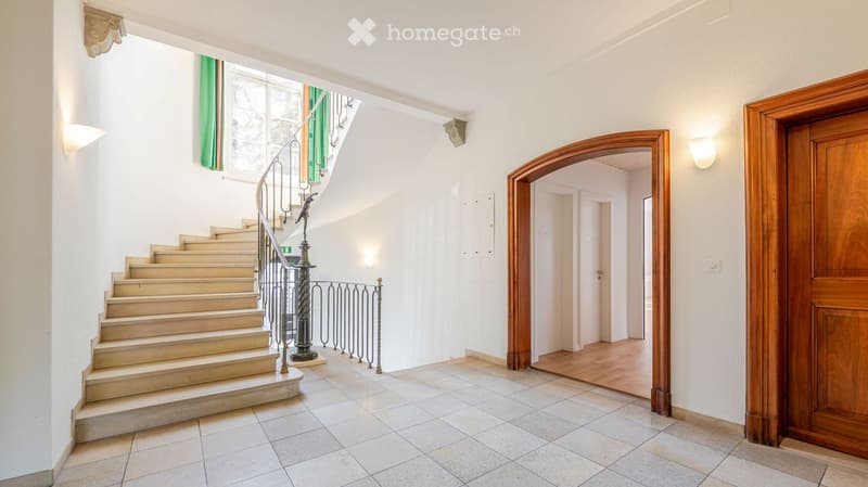 Spacious hallway with a curved staircase leading upwards, featuring a metal railing and a decorative lamp post. The hallway has tiled flooring, white walls, and a wooden door on the right side.