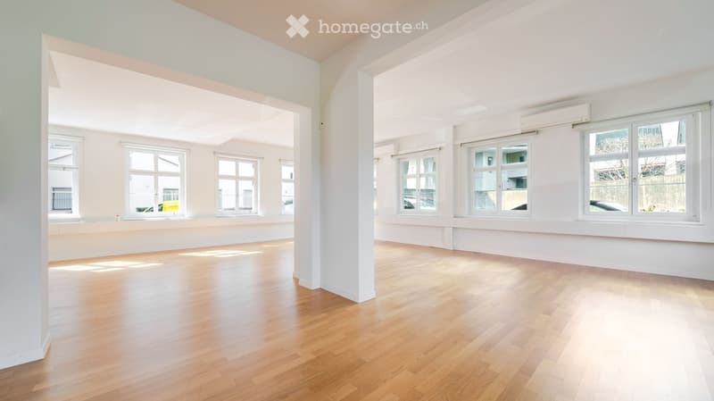 Empty room with light wooden flooring, white walls, and multiple large windows allowing natural light to fill the space.