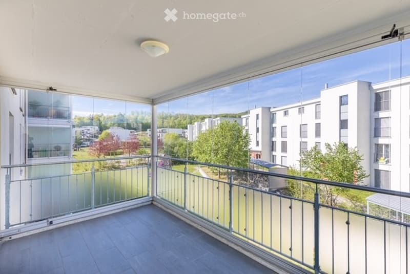 Spacious balcony with modern glass railings, tiled floor, and expansive views of a green park and surrounding buildings.