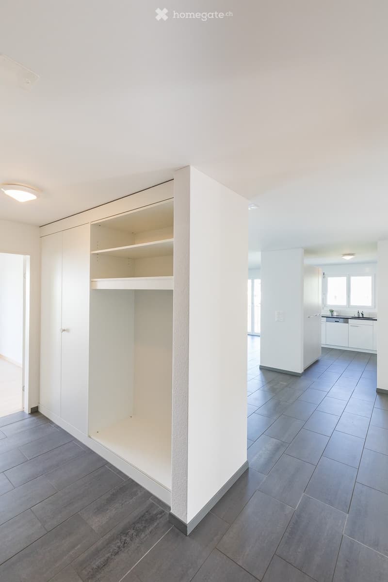 Empty room with grey tiled flooring, white walls, built-in shelving unit, and a small adjacent kitchen area with white cabinets and a sink.