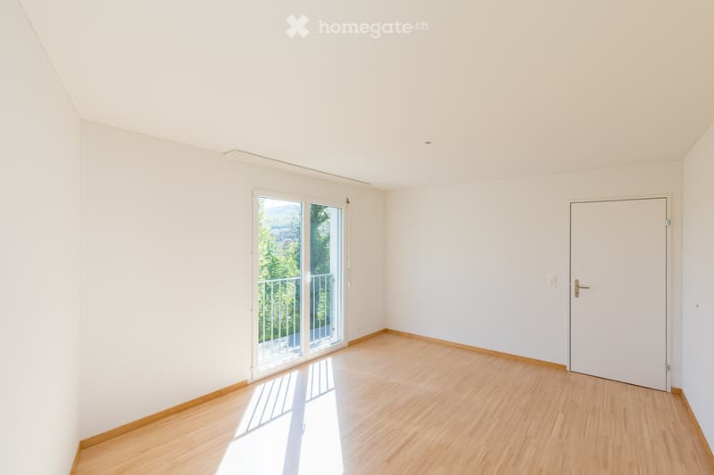 Empty room with light wooden flooring, white walls, and a large sliding glass door leading to a balcony with a view of greenery.