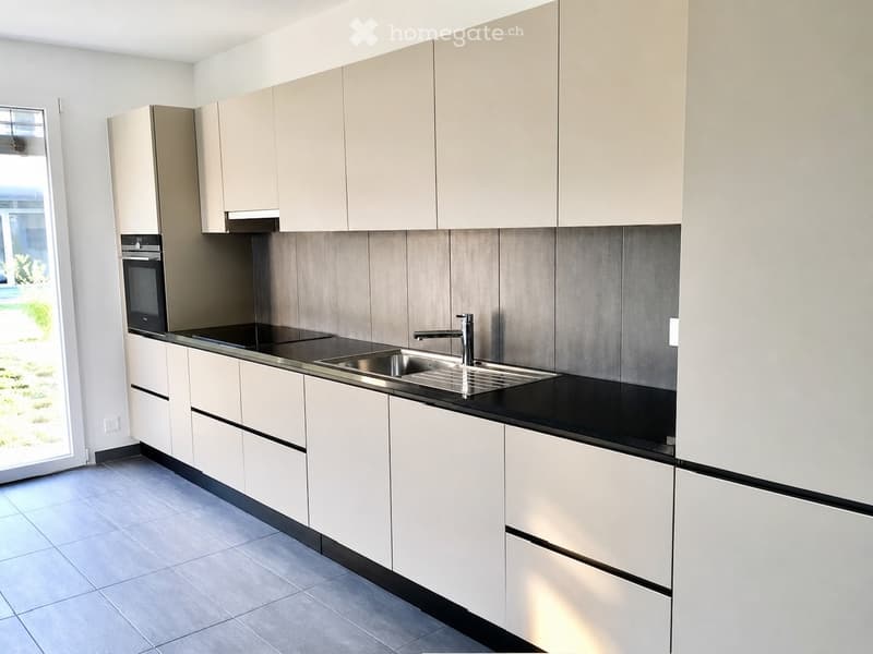 Modern kitchen with light-colored cabinets, stainless steel sink, and built-in oven. The countertops are black, and the backsplash features a textured grey tile design.
