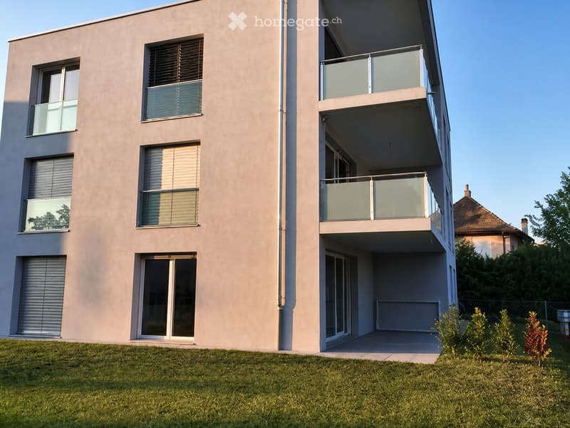 Modern two-story building with beige facade, multiple windows with blinds, and two balconies with glass railings.