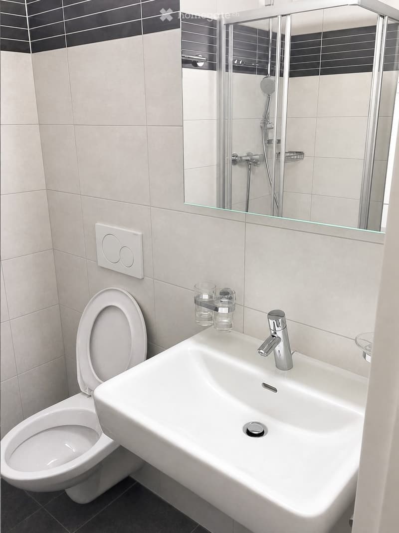 Modern bathroom with a white toilet, sink, and shower area. The sink has a chrome faucet and two cups on the counter. A large mirror is above the sink, and the walls are tiled in light gray with black accents.