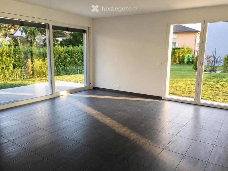 Empty room with dark tiled flooring, large sliding glass doors leading to a garden, white walls, and natural light streaming in.