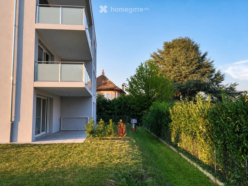 Well-maintained garden with green grass, trimmed bushes, and a modern building with balconies in the background.