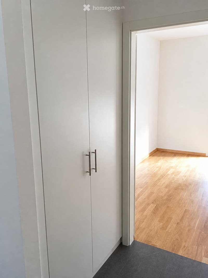 A modern, minimalist hallway with white walls, dark grey tiled flooring, and a wooden floor visible in the adjacent room. The hallway features a white cabinet with a silver handle on the left side.