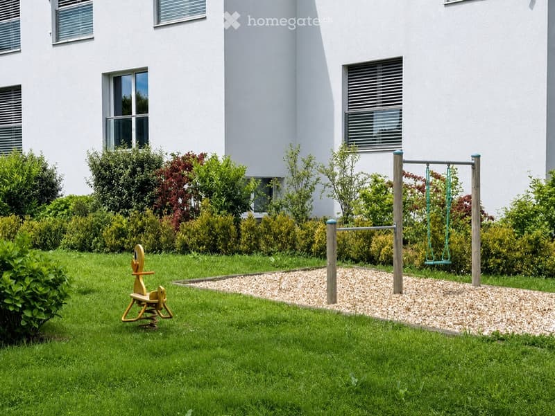 Well-maintained garden with a swing set, a rocking horse, and neatly trimmed bushes surrounding a modern white building.