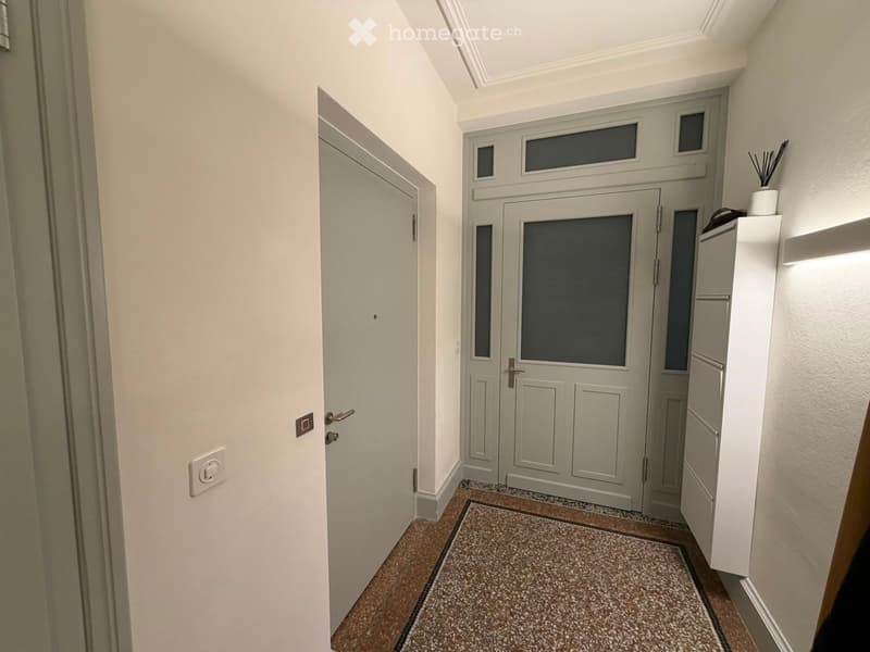 A narrow hallway with a patterned rug, white walls, and a double door at the end. There is a small shelf with decorative items on the right side.