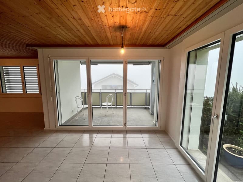 Bright, tiled indoor space with large sliding glass doors leading to a balcony area featuring two white chairs and a small table.