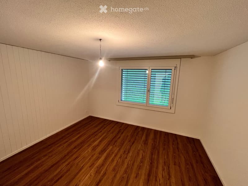 Empty room with wooden flooring, white walls, and a single window with blinds. A single light fixture hangs from the ceiling.