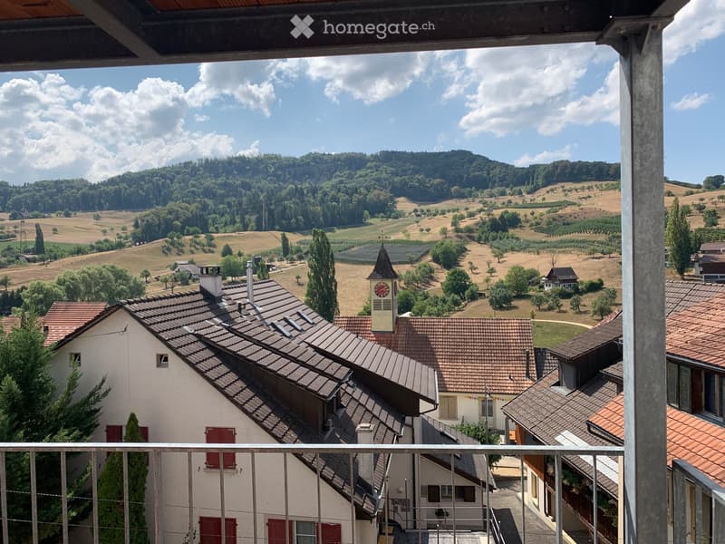 Balcony overlooking a scenic countryside with rolling hills, scattered trees, and a small village with a church tower.