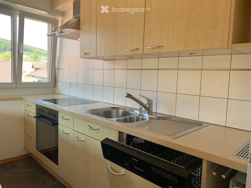 Modern kitchen with light wood cabinets, stainless steel sink, oven, and dishwasher. Features a large window with a scenic view.