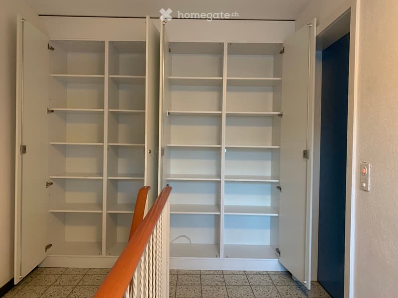 Empty room with white built-in shelving unit, tiled floor, and a small section of a wooden chair visible in the foreground.