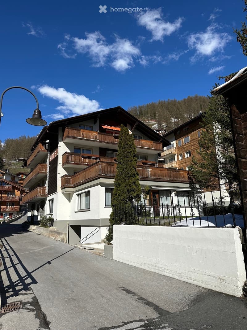 Multi-story building with balconies, wooden railings, and a stone facade, surrounded by trees and a clear blue sky.
