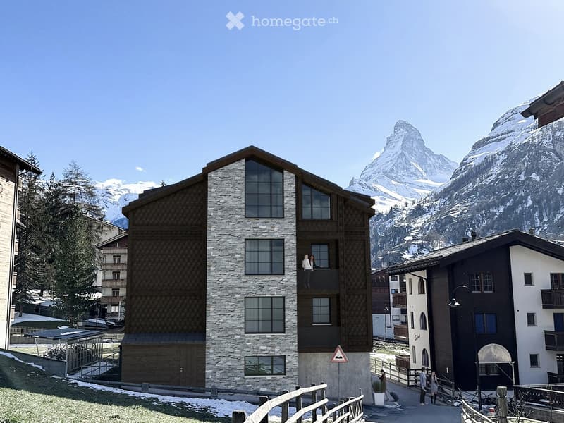 A modern stone and wood building with large windows and balconies, situated in a picturesque alpine village with snow-capped mountains in the background.