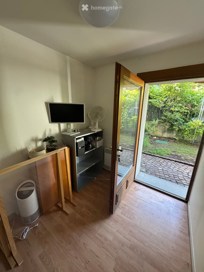 A small, well-lit room with wooden flooring, a screen on a cabinet, a fan, a potted plant, and an open door leading to a garden area.