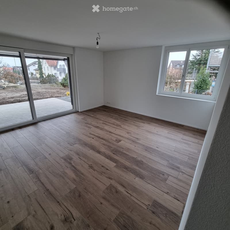 Empty room with wooden flooring, large windows, and a sliding glass door leading to a patio.